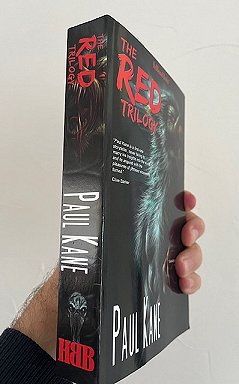 photograph of a man's hand holding up a copy of The Red Trilogy by Paul Kane, so that the spine of the book is to the fornt of the picture