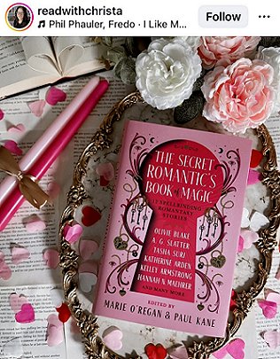 screenshot from @readwithcrista. Photograph of a copy of The Secret Romantic's Book of Magic, edited by Marie O'Regan and Paul Kane, lying on a round gilt frame covered in pink and white confetti, on top of loose book pages. To the left are two pink candles tied with a gold ribbon, and pink and white roses are at the top of the picture