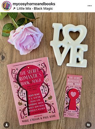 photograph of a copy of The Secret Romantic's Book of Magic, edited by Marie O'Regan and Paul Kane, on a wooden surface. To the right of the book is a Secret Romantic's bookmark, and at the top of the picture is a pink rose to the left, and a white ceramic ornament that spells LOVE on the right