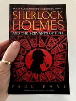 Man's hand holding a copy of Sherlock Holmes and the Servants of Hell, by Paul Kane