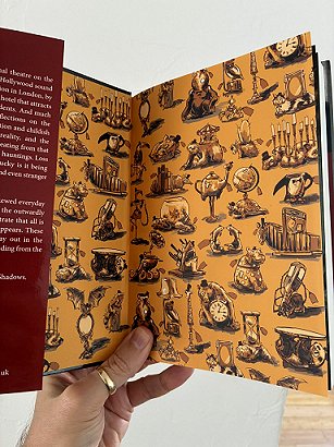 photograph showing a hand holding up a copy of Shadowplays, edited by Peter Coleborn and Mike Chinn, to show the orange endpapers decorated with clocks, candlesticks, talbes, bears and various other items