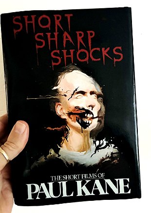 photograph showing a man's hand holding up a copy of Short Sharp Shocks by Paul Kane