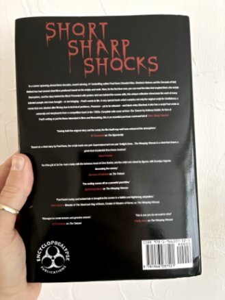 photograph of a man's hand holding up a copy of Short Sharp Shocks by Paul Kane, to show the back cover