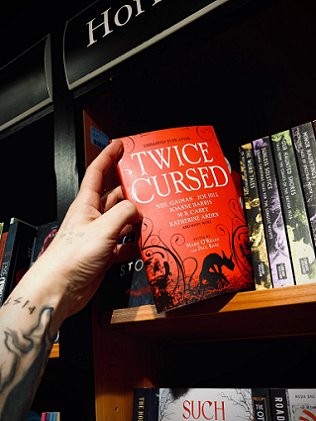 Image of a man's hand and tattooed arm taking a copy of the book Twice Cursed, edited by Marie O'Regan and Paul Kane, off a bookshelf