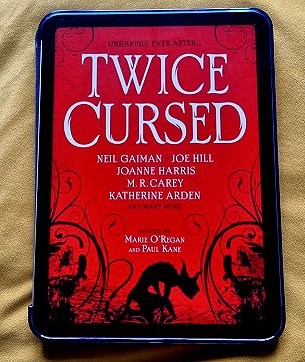 Picture of a Kindle laying on a gold cloth. Kindle features the cover image of Twice Cursed, edited by Marie O'Regan and Paul Kane