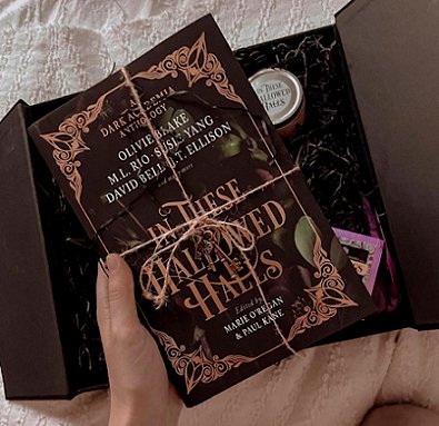 image of a woman's hand holding a copy of In These Hallowed Halls, edited by Marie O'Regan and Paul Kane, above a black box containing black tissue paper, an In These Hallowed Halls candle and an Earl Grey teabag - the box is lying on a cream fleece cloth