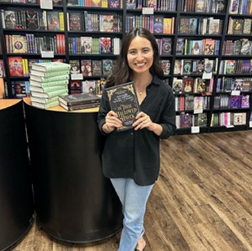 photograph showing author Olivie Blake standing in front of bookshelves, beside a pile of books on a table, holding a copy of In These Hallowed Halls, edited by Marie O'Regan and Paul Kane
