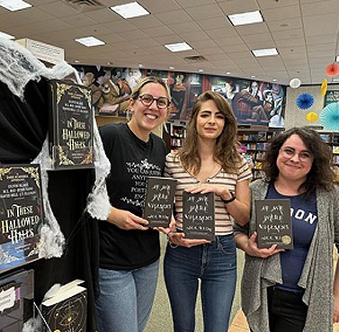 photograph showing M L Rio standing in the middle of a blonde woman and a woman with dark hair, all holding copies of M L Rio's novel If We Were Villains. There are copies of In These Hallowed Halls, edited by Marie O'Regan and Paul Kane, on the display beside them
